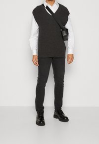 Chaleco de punto gris sobre una camisa blanca, combinado con pantalones negros ajustados y botas negras con cordones. Incluye un pequeño bolso cruzado negro.