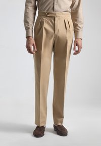 Pantalones de pliegues beige de tela suave, corte ajustado, con cierre de botón en la cintura, combinados con calzado marrón oscuro.