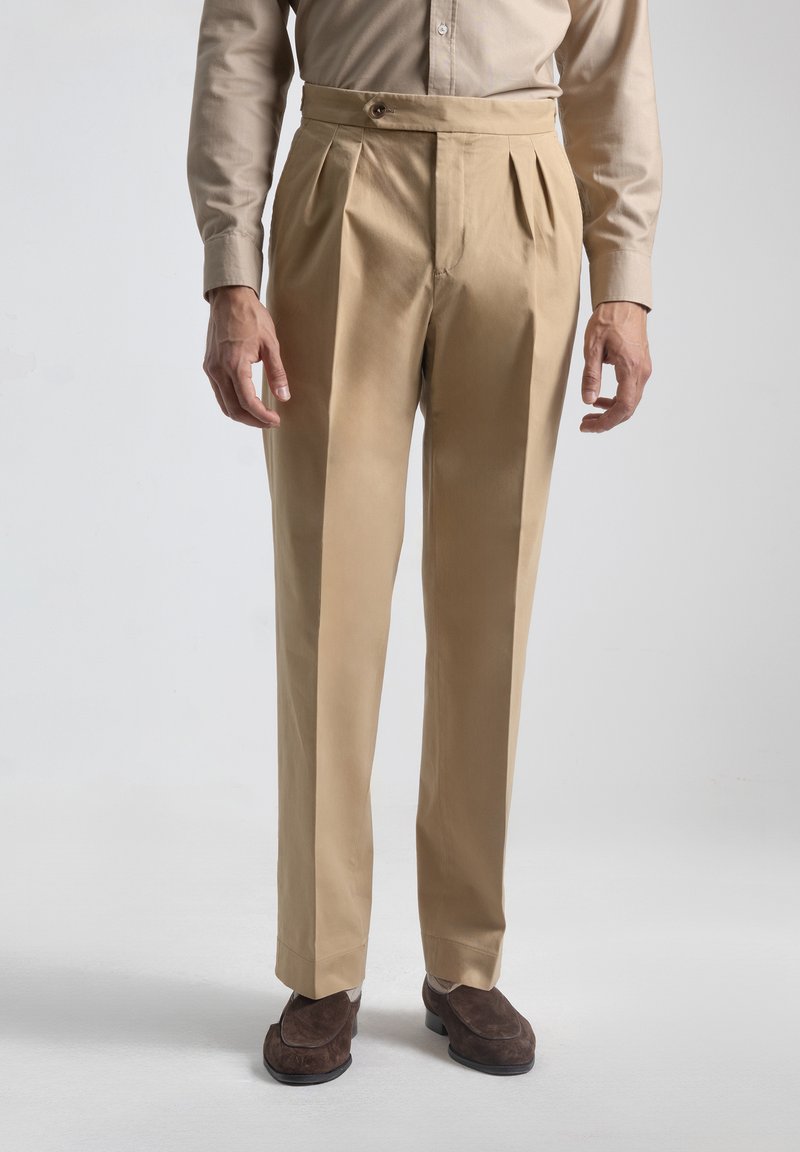 Pantalones de pliegues beige de tela suave, corte ajustado, con cierre de botón en la cintura, combinados con calzado marrón oscuro.