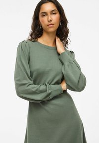 Vestito verde lavorato a maglia con maniche lunghe e morbide a sbuffo e scollatura rotonda. Tessuto morbido e corpetto aderente, leggermente svasato in vita.