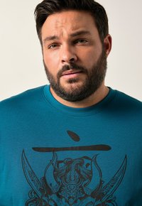Camiseta de algodón en color aguamarina con un gran diseño gráfico negro que presenta patrones intrincados, incluyendo un rostro estilizado y acentos de espada.