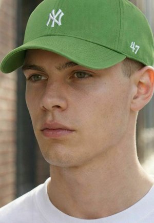 Junger Mann mit hellem Teint, der eine grüne Baseballmütze mit weißem NY-Logo und ein weißes Hemd trägt, schaut im Freien leicht zur Seite.