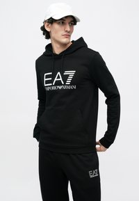 Fekete pamut kapucnis pulóver kenguruzsebbel, fehér "EA7 Emporio Armani" logóval. Fekete nadrággal párosítva, egységes megjelenést teremtve.