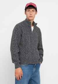 Suéter gris de punto con cuello alto y media cremallera, que presenta una tela texturizada y puños acanalados, combinado con jeans azules claros y una gorra rosa.