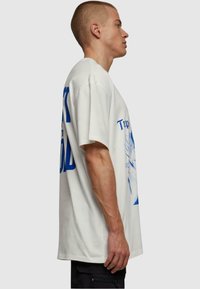 Hvid oversized T-shirt lavet af bomuld, med blå tekst og grafisk design på ryggen. Korte ærmer, afslappet pasform, set fra siden.