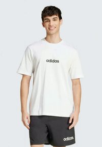 Hvit t-skjorte med korte ermer som har en svart "adidas"-logo sentrert på brystet. Glatt tekstur, ledig passform, kombinert med svarte shorts.