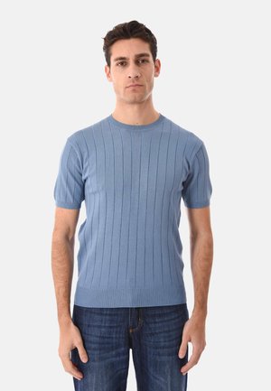 Uomo che indossa un maglione aderente a maniche corte blu a coste e jeans blu scuro, in piedi davanti a uno sfondo chiaro e uniforme.