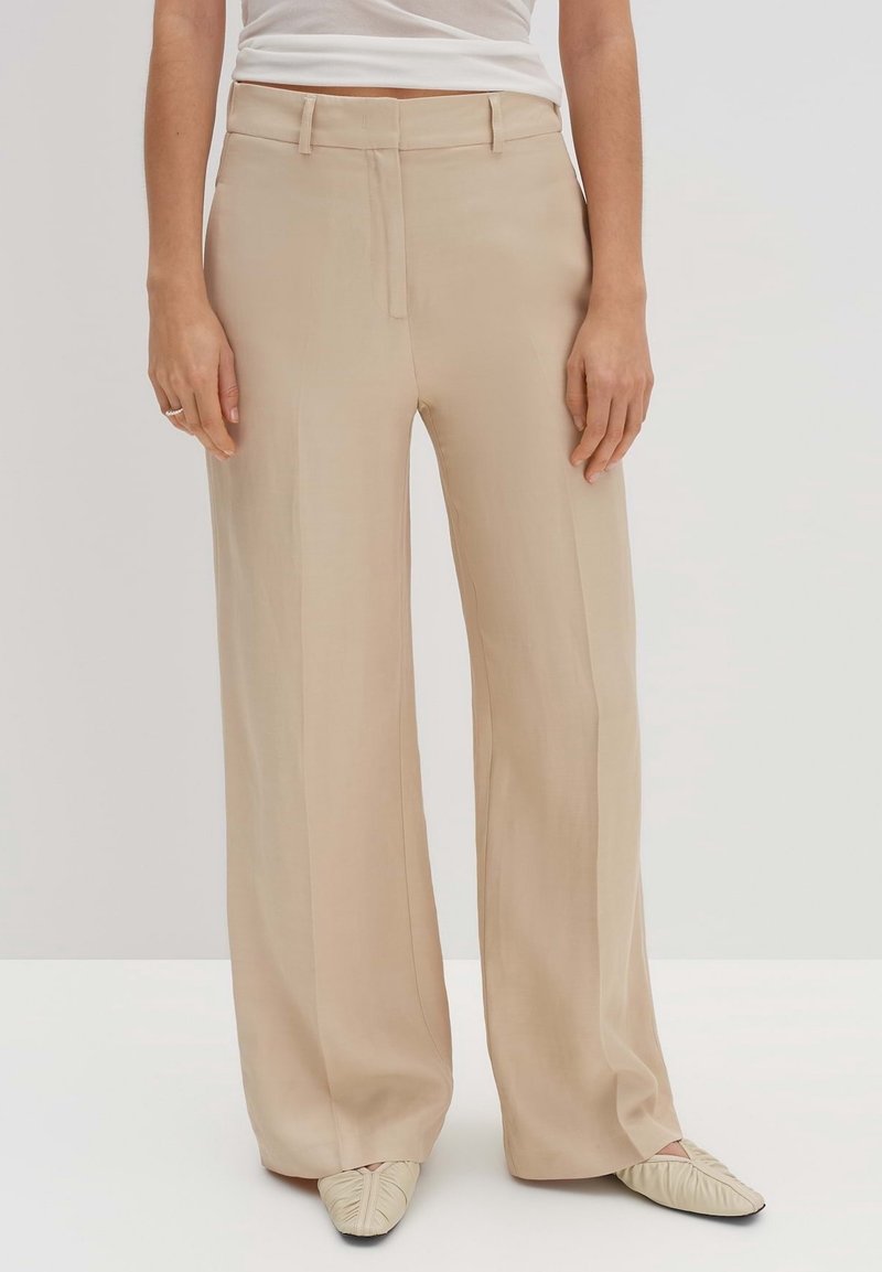 Femme portant un pantalon large beige, des chaussures plates crème froncées, et un haut blanc, debout devant un fond clair uni.