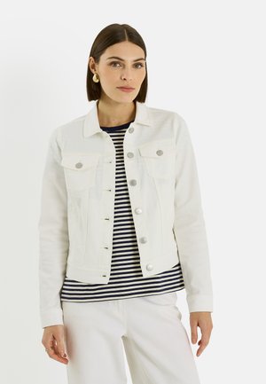 Femme portant une veste en jean blanche sur un chemisier rayé bleu marine et blanc, assortie à un pantalon blanc, sur un fond blanc uni.