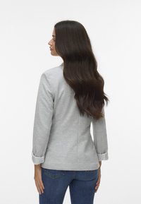 Szara bluzka z długim rękawem i podwiniętymi mankietami, dopasowany krój oraz gładka tekstura, zestawiona z ciemnoniebieskimi dżinsami. Widok z tyłu odzieży.