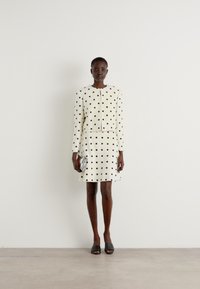 White dress with black polka dots, knee-length, round neckline, long sleeves, and a fitted bodice. Model holds a small silver clutch.