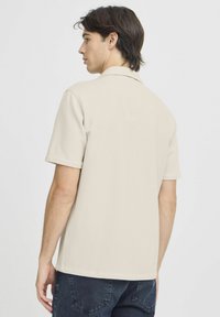 Camisa polo de manga corta beige con un patrón acanalado texturizado, corte relajado y un cuello plano, combinada con jeans oscuros.