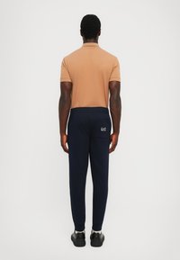 Brown short-sleeve polo shirt and navy sweatpants with gathered cuffs and logo detail, viewed from the back, worn with black shoes.