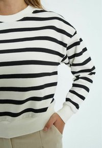 Sivi dukserica s crno-belim prugama koja ima kružni izrez, rebraste rukave i donji rub, te opušten kroj. Naglasak na mekoj teksturi materijala.