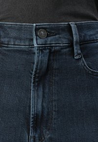Blugi din denim de culoare bleumarin închise, cu un fermoar tradițional, bucle pentru curea și detalii de cusătură discrete de-a lungul cusăturilor și buzunarelor.