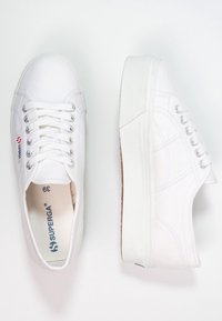 Baskets en toile blanche avec semelle plate, bout arrondi, œillets argentés, semelle extérieure en caoutchouc et logo de la marque discret sur le côté.