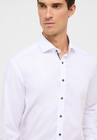 Camicia bianca con bottoni, realizzata in un tessuto testurizzato, colletto interno a fantasia a contrasto e bottoni marrone scuro sul davanti.