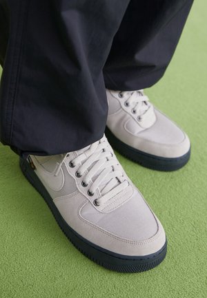 Baskets gris clair avec semelles noires portées avec un pantalon foncé, debout sur un sol recouvert de tapis vert.
