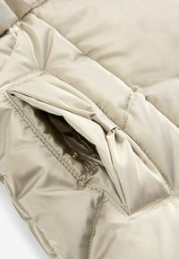 Sección de chaqueta acolchada de puffer beige con un bolsillo inclinado. Tejido suave y brillante con sutiles detalles de costura y cierre a presión.