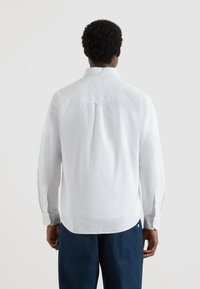 Uomo con capelli ricci corti che indossa una camicia bianca a maniche lunghe e pantaloni blu scuro, di spalle contro uno sfondo neutro.