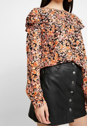 Kvinde iført sort blomsterlangærmet bluse med orange blomster og sort knapdet lædermininederdel, stående mod en ensfarvet baggrund.