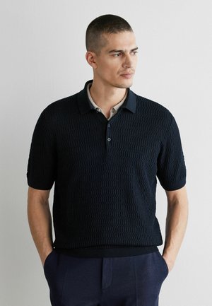 Mann mit kurzen Haaren trägt ein strukturiertes schwarzes kurzärmeliges Poloshirt und marineblaue Hose, steht mit den Händen in den Taschen und schaut nach rechts an einer weißen Wand.