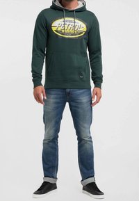 Felpa verde con logo grafico, cappuccio interno grigio, tasca a marsupio, jeans blu usurati e sneaker nere. Vestibilità casual, materiale in cotone.