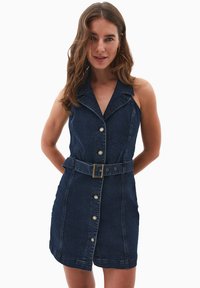 Mörk denimklänning med figursydd midja, ärmlös design, krage, knäppning med knappar och ett bälte. Har synlig sömnad och fem knappar.