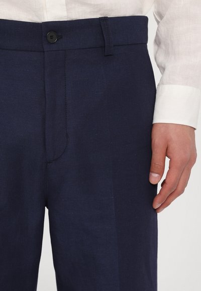 Sötétkék vászon nadrág gombos záródással, testhezálló fazonnal és finom textúrával. A derékpántra nyúló kéz kiemeli a dizájn részletet.