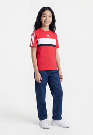 Fille souriante debout portant un t-shirt rouge à manches courtes avec des rayures blanches et noires, un jean bleu et des baskets blanches sur fond blanc.