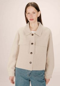 Kvinde med langt brunt hår, iført en beige knapjakke med klaplommer og blå jeans mod en ensfarvet lys baggrund.