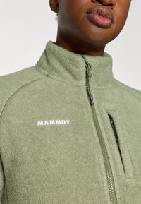 Grüne Fleecejacke mit hohem Kragen, durchgehendem Reißverschluss und einer Fronttasche. Merkt sich das weiße "MAMMUT"-Logo auf der Brust. Weiche Textur, lässiges Design.