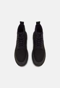 Para czarnych sneakersów typu high-top z teksturowanej tkaniny, zaokrąglonymi noskami, grubymi podeszwami i uchwytami, pokazana z góry na białym tle.