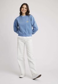 Sweatshirt en coton bleu avec un petit graphique de soleil, assorti à un pantalon évasé blanc à rayures et des baskets blanches. Texture lisse, design décontracté.