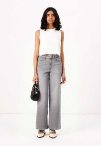 Top corto bianco, jeans grigi a vita alta e gamba larga, borsa nera e scarpe bicolore con punta affusolata; tessuto liscio e linee pulite.