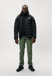 Chaqueta de plumas negra con cremallera, llevada sobre una camisa negra; pantalones cargo verdes con bolsillos y zapatos de senderismo negros. Mochila ajustable.