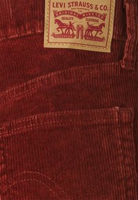 Tissu en velours côtelé rouge avec des rainures verticales, comprenant une étiquette beige avec des graphiques de cheval et d'homme. Comprend des surpiqûres et plusieurs poches.