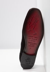 Zwarte suède schoenzool met een rode binnenkant waarop een skelet speelt op een saxofon en gegraveerde tekst: "Zonder muziek zou het leven een fout zijn."