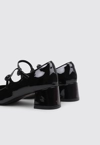 Chaussures Mary Jane en cuir verni noir avec des talons carrés arrondis et des fermetures à bouton à double sangle sur un fond gris clair.