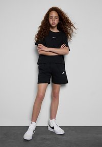 Svart Nike t-skjorte og shorts med en hvit logo. Modellen har på seg hvite høyhælte joggesko. Bakgrunnen er en ensfarget grå vegg.