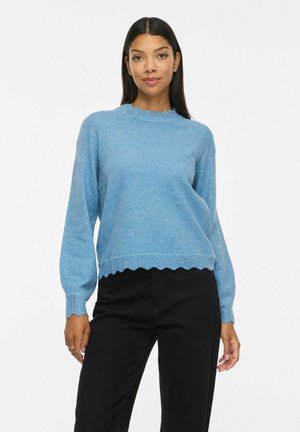 Femme portant un pull bleu clair à bords festonnés et un pantalon noir, debout sur un fond blanc uni.