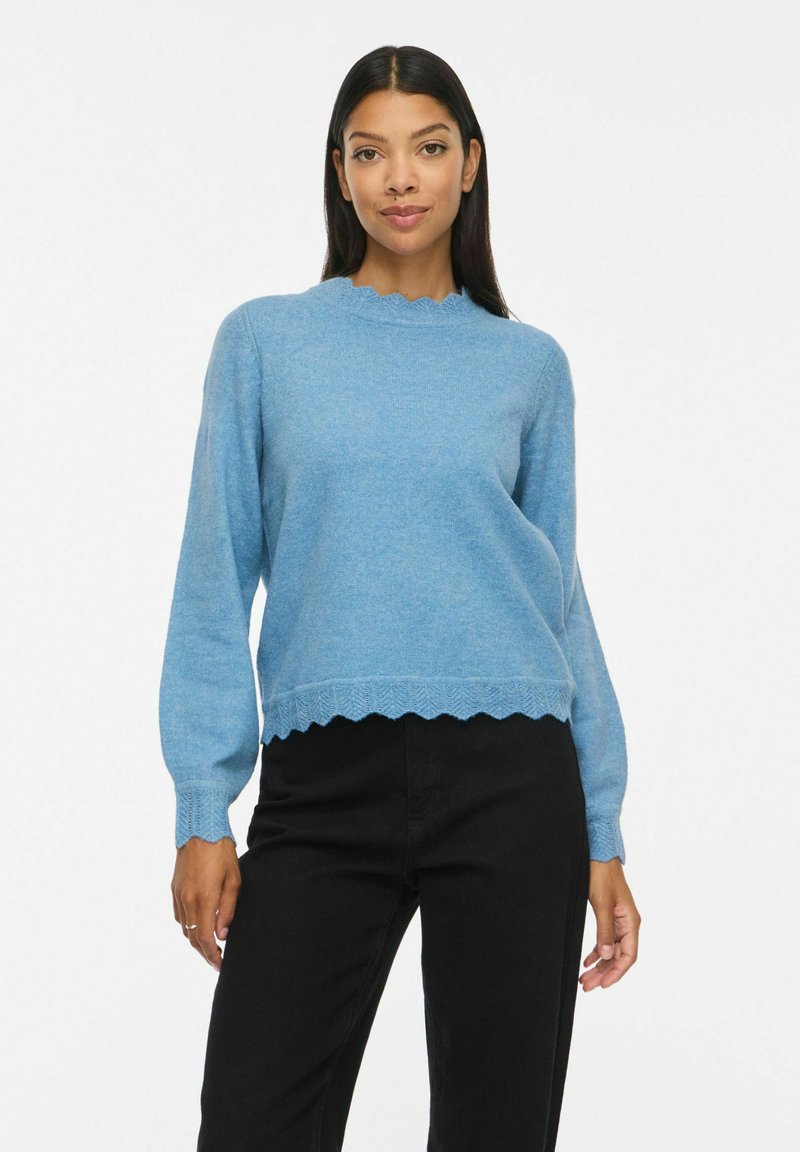 Femme portant un pull bleu clair à bords festonnés et un pantalon noir, debout sur un fond blanc uni.