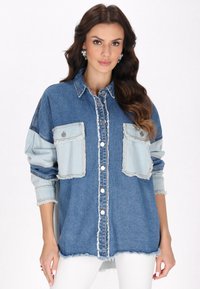 Lichtblauwe spijkerblouse met twee grote borstzakken, versleten zoom, knoopsluiting en lange mouwen in een contrasterend donkerder blauw.