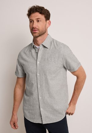 Mies, jolla on lyhyet kiharat ruskeat hiukset, pukeutuneena vaaleanharmaaseen lyhythihaiseen napilliseen paitaan ja tummiin housuihin yksivärisellä taustalla.