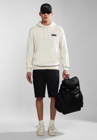 Sudadera blanca con bolsillo frontal, pantalones cortos de carga negros, zapatillas crema y gorra negra. Mochila negra con parte superior enrollable y logo. Lentes de sol puestos.