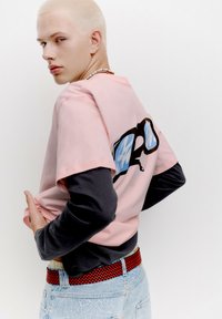 Tricou grafic roz cu mâneci negre, prezentând un grafic cu ochelari de soare albi și negri, asociat cu blugi deschiși la culoare și o curea roșie și neagră.