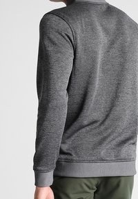 Hombre vistiendo un suéter de manga larga gris ajustado con puños y dobladillo acanalados, combinado con pantalones verde oliva, visto desde atrás.
