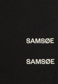 Juodas audinys su pakartotiniu baltu tekstu "SAMSOE", kur antrame variante perbraukta "Ø". Lygi tekstūra, minimalistinis dizainas.