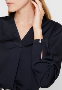 Marine blouse met een V-hals, geplooide details en lange mouwen met knopaccenten. Glad materiaal met een zachte glans.