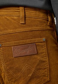 Corduroy broek in een warme, goudbruine kleur met een bruine leren Wrangler-label, voorzien van subtiele stikselaccenten en details op de zakken.